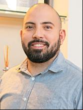 Homem sorridente com barba, usando camisa social cinza clara, posando em ambiente corporativo iluminado.